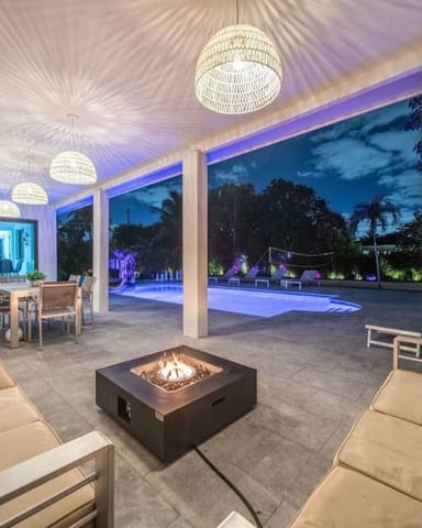 Modern covered patio with fire pit, lounge seating, pendant lights, and views of illuminated pool and tropical landscape at dusk