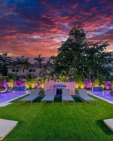 Luxury backyard at sunset with two illuminated pools, manicured lawn, lounge chairs, and colorful landscape lighting.