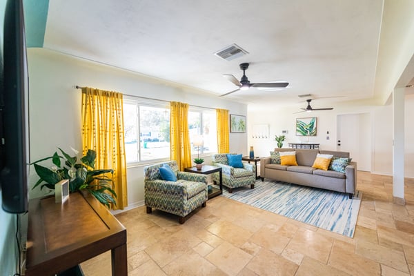Bright open-plan living room with yellow curtains, patterned armchairs, gray sofa, tile and wood flooring, and large windows overlooking a parking area.