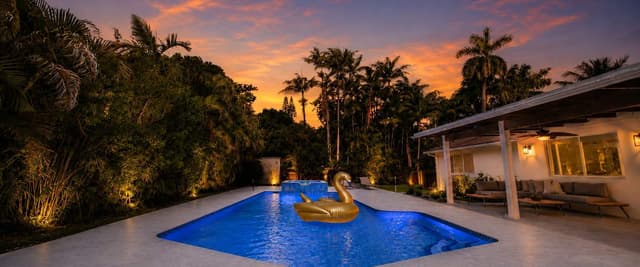 Backyard pool at dusk with gold swan float, palm trees, modern white house, and colorful sunset sky