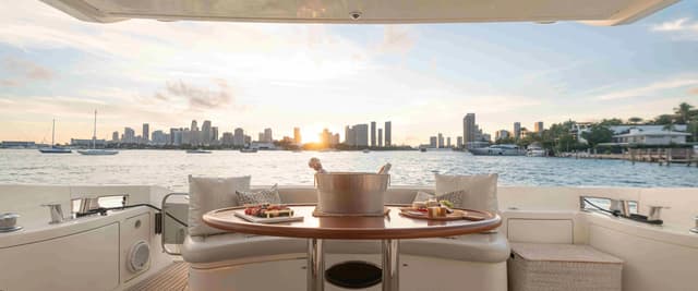 Luxury yacht flybridge with dining table, city skyline and sailboats visible across the water at sunset