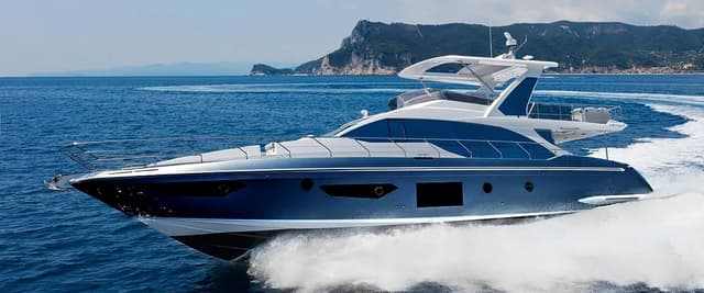 Luxury motor yacht with blue and white hull in shallow coastal waters, rocky island and seaside town visible in background