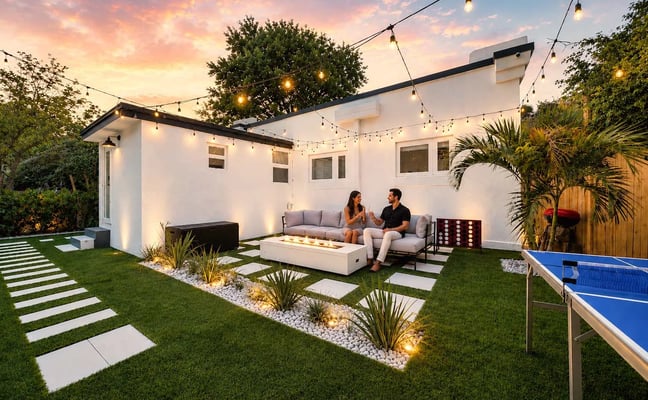Modern white backyard house with couple sitting on gray sofa by fire pit, string lights overhead, palm plants, and ping pong table at dusk.